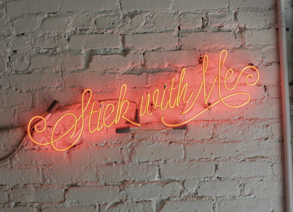 Photo of a neon sign that says, in fiery yellow-orange, "Stick with Me." It's up against a whitewashed brick wall and casting a red glow against the stone. Photo by Cory Bouthillette @ Unsplash.